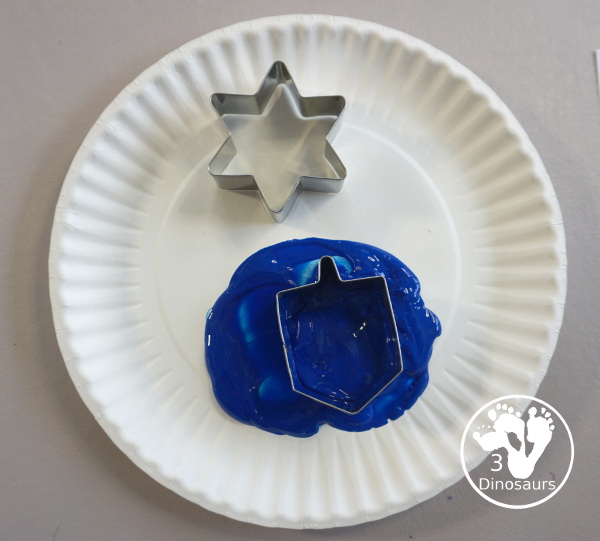  Hanukkah Stamping Painting with Cookie Cutters is a great Hanukkah craft you can do with different ages. And a fun mix of Hanukkah themes you can find in the cookie cutters.